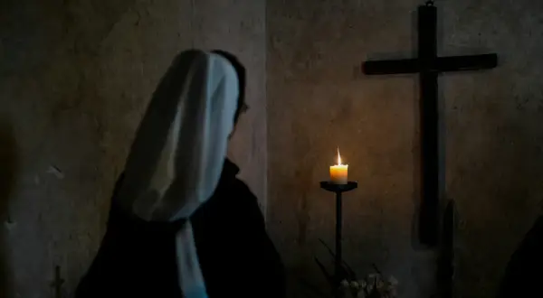 Zakonnica w białym habicie modli się przed krzyżem i płonącą świecą.