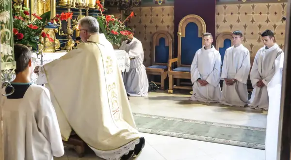 Ksiądz w szatach odprawia nabożeństwo, klęczą ministranci w białych albach.