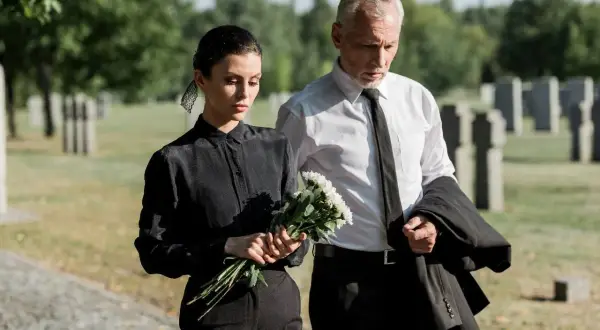 Para idąca przez cmentarz, kobieta w czarnej sukience trzyma bukiet białych kwiatów, mężczyzna w białej koszuli i czarnym krawacie niesie marynarkę.